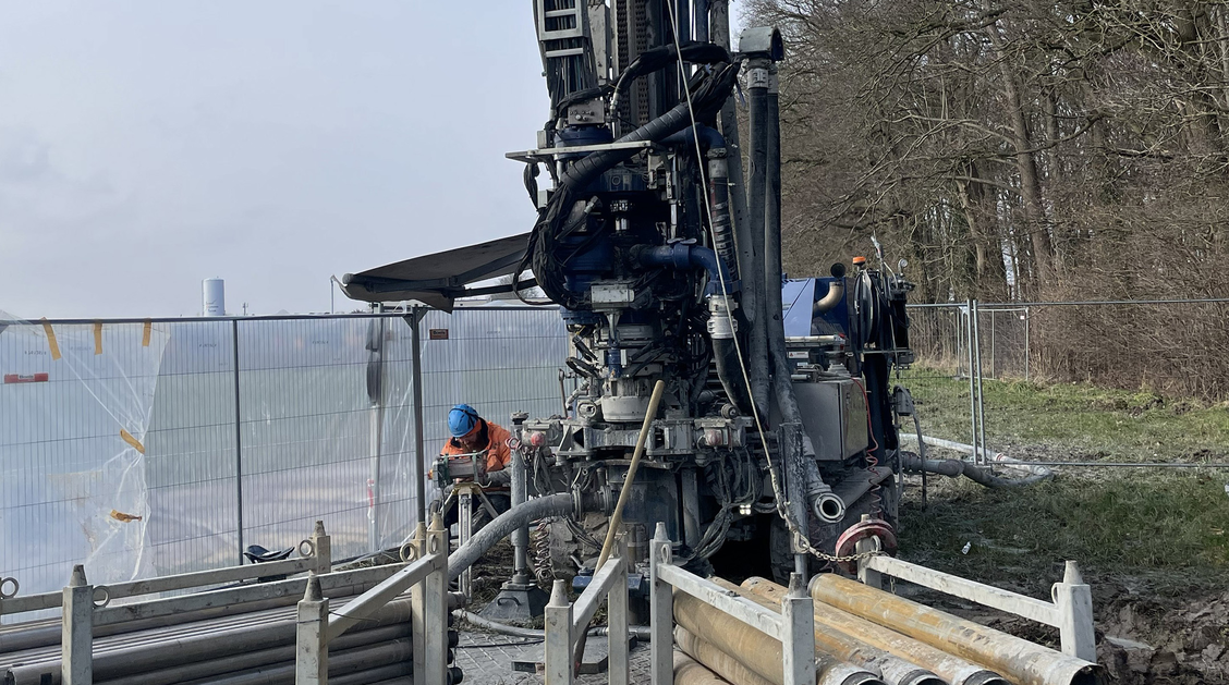 Das Bild zeigt eine Bohranlage auf einer ländlich anmutenden Fläche, links von der Anlage ist ein Bauzaun zu sehen. 