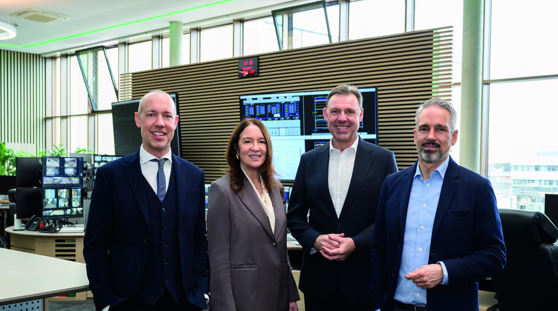 Das Bild zeigt vier Personen in einem großen Raum mit Monitoren im Hintergrund. 