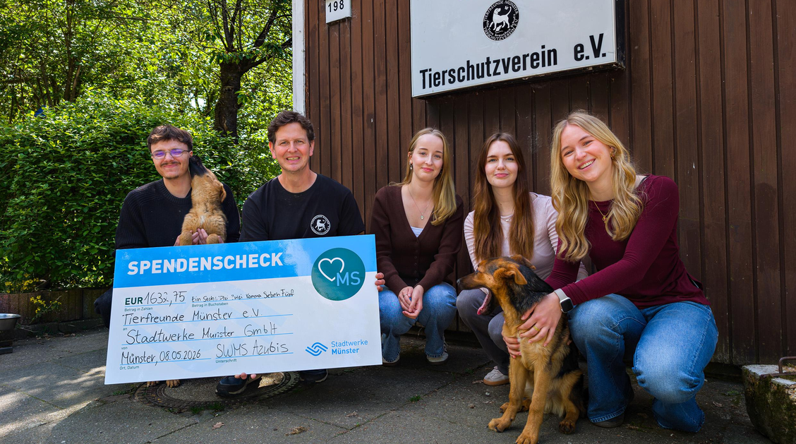 Fünf Personen und zwei Hunde hocken vor einer Holzwand, an das ein Schild des Tierschutzvereins angebracht ist. Eine Person hält einen übergroßen Spendenscheck. 