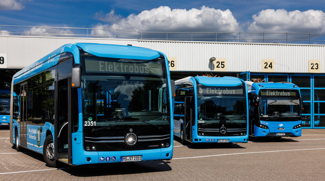 Drei Busse stehen vor einer großen Halle, ihre Zielanzeige zeigt "Elektrobus"