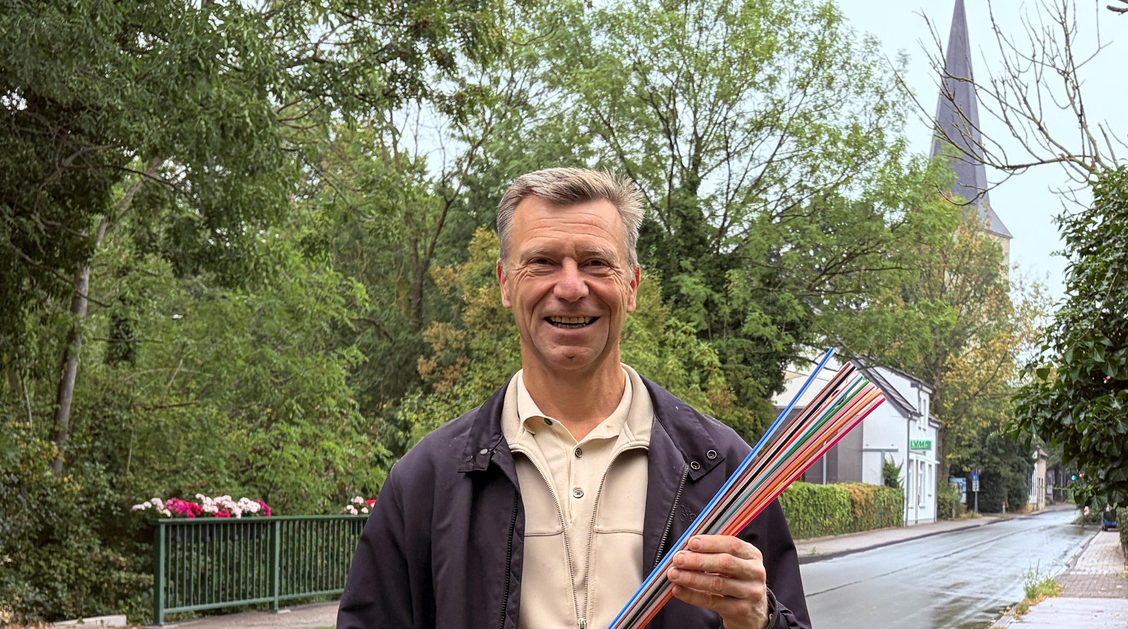 Ein Mann steht an einer Straße und hält ein buntes Kabelbündel in der Hand. Im Hintergrund ist ein Kirchturm, eine Straße und ein Brückengeländer mit Blumenkästen zu sehen. 