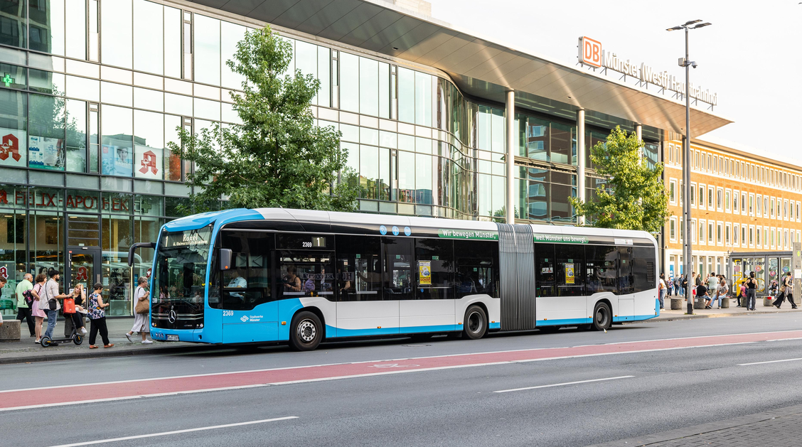 Ein Bus steht vor einem Gebäude an einer Haltestelle, es eilen Menschen vorbei 