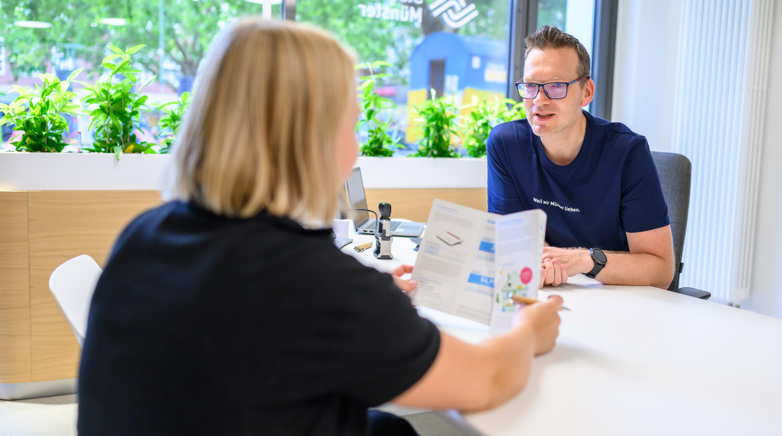 Zwei Personen sitzen an einem Schreibtisch. Von einer Person sieht man nur den Hinterkopf. Sie hält einen Flyer. Die andere Person sitzt ihr gegenüber und ist ihr zugewandt.
