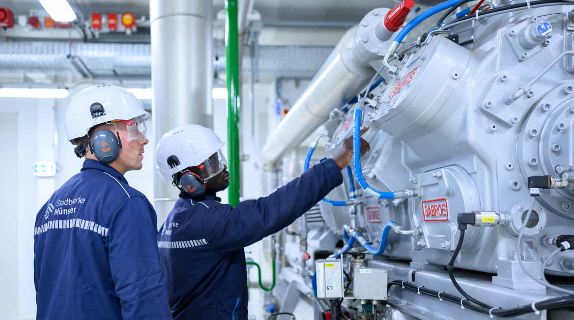 Zwei Personen in Schutzausrüstung stehen in einem hellen Raum an einer technischen Anlage. 
