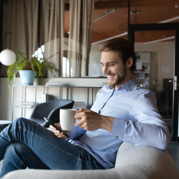 Ein junger Mann hat einen Kaffee und sein Smartphone in der Hand und sitzt auf einem Sessel.
