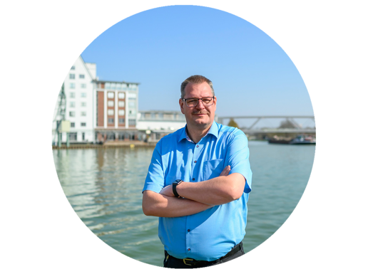 Ein Mann steht mit verschränkten Armen am Hafen und lächelt.