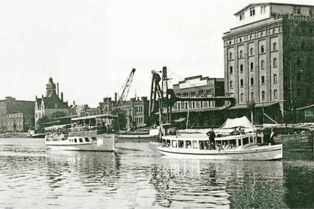 Zwei weiße Ausflugsschiffe verlassen den Hafen von Münster. Im Hintergrund sind Industrie- und Lagergebäude zu sehen
