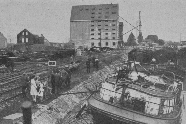 Ein beschädigtes Schiff liegt im nur noch halb mit Wasser gefüllten Hafenbecken, am Ufer sind zerstörte Gebäude zu sehen