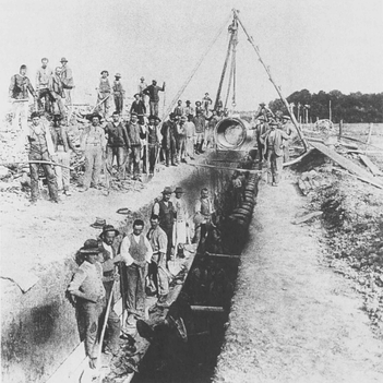 In einen Graben wird ein Wasserrohr verlegt. Viele Menschen arbeiten daran