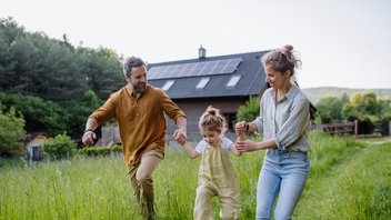 Eine Familie läuft glücklich durch ein Feld, im Hintergrund ist ein Haus mit einem Solarpanel auf dem Dach zu sehen.