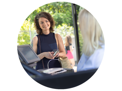 Eine Frau mittleren Alters steht lächelnd in der Bustür. Die Busfahrerin ist von hinten zu erkennen.