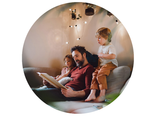 Ein Vater sitzt mit seinen beiden kleinen Kinder auf einem Sofa und liest aus einem Buch vor.