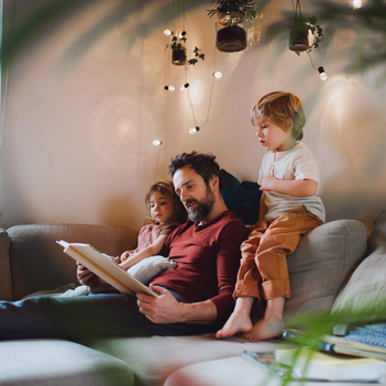 Ein Vater sitzt mit seinen beiden kleinen Kinder auf einem Sofa und liest aus einem Buch vor.