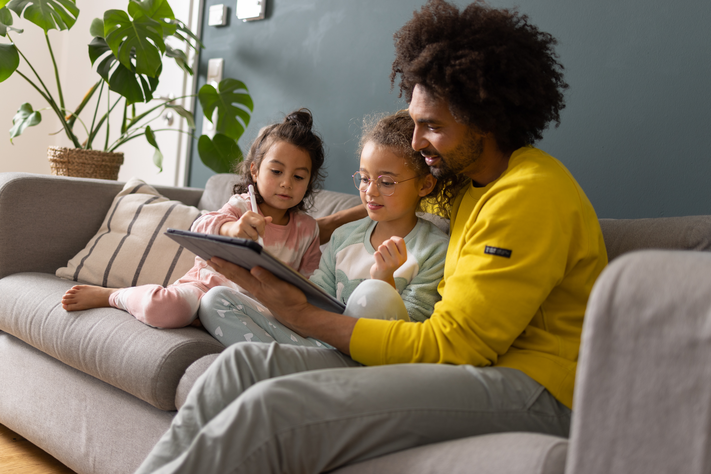 Vater sitzt mit seinen zwei kleinen Kindern auf dem Sofa und schauen auf das Tablet.