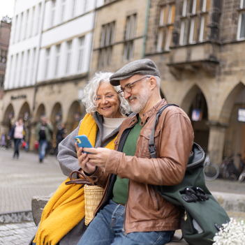 Eine älteres Ehepaar sitzt an einem Brunnen und schaut gemeinsam auf ein Smartphone.