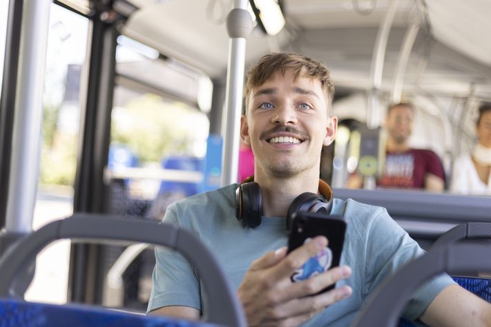 Ein junger Mann sitzt mit Kopfhörern um seinen Hals und seinem Smartphone in der Hand in einem Stadtwerke-Bus.