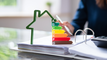 Ein Plastikhaus mit den Energieeffizienzklassen A bis G steht auf einem aufgeschlagenen Ordner. Daneben liegt ein Taschenrechner.