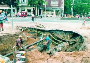 Ein großes Loch an einer Straßenecke mit vielen Rohrleitungen und zwei Bauarbeitern in der Grube. 